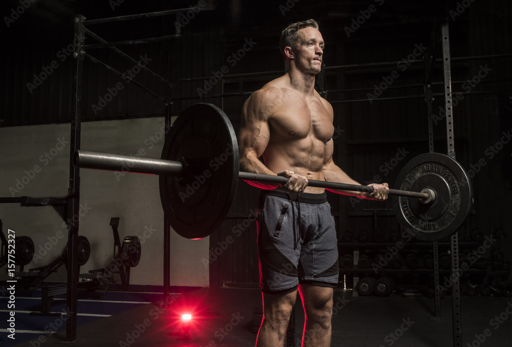 Muscular White Caucasian man doing a   barbell bicep curl  in a dark grungy gym wearing shorts and showing muscular body and six pack abs 