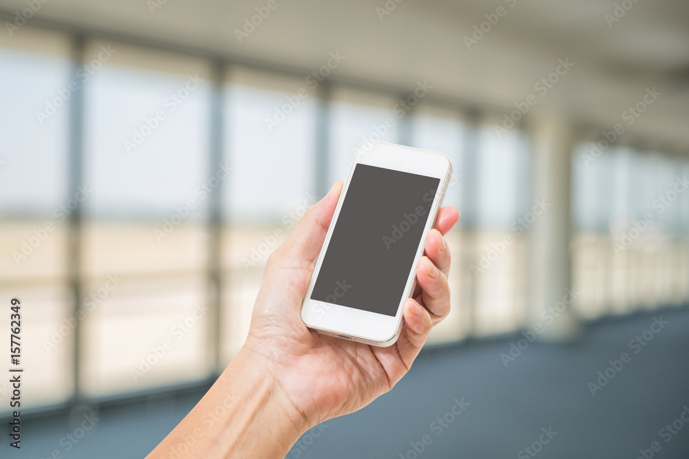Hand holding blank screen mobile phone with abstract blurred image of office lobby space interior view background. Hand holding blank screen mobile phone with abstract blurred image of office lobby space interior view background.