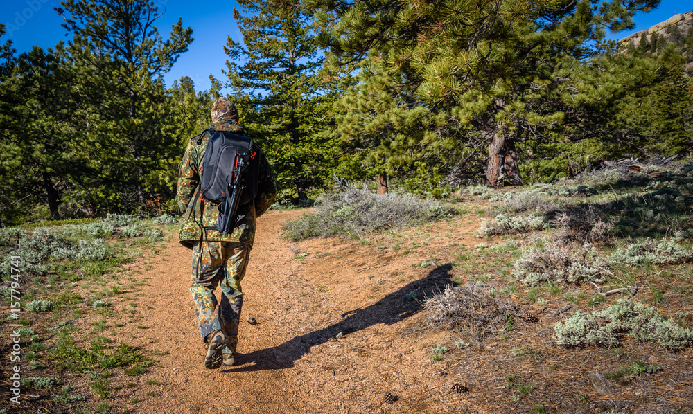 Naklejka premium A man photographer hiking in camouflage outfit discovering nature in the forest with DSLR photo camera, lenses, tripod in the backpack. Travel photography lifestyle concept. Rear view