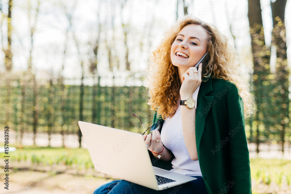 Portrait of beautiful girl having fluffy blonde hair holding smartphone in one hand and sunglasses in other having telephone conversation being excited to speak with her best friend using laptop Portrait of beautiful girl having fluffy blonde hair holding smartphone in one hand and sunglasses in other having telephone conversation being excited to speak with her best friend using laptop