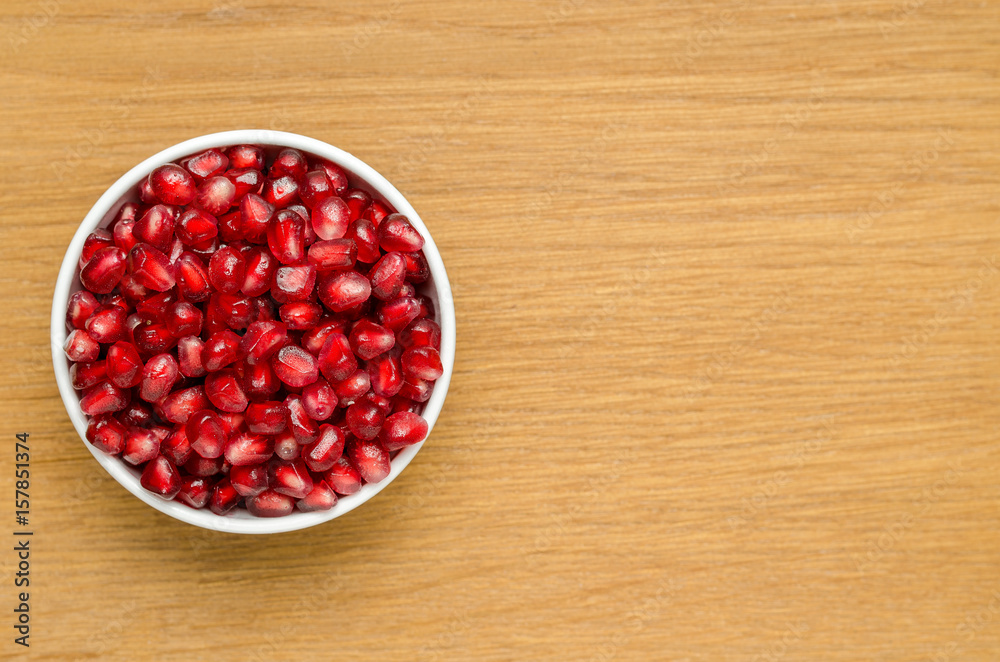 Grains Of Pomegranate In A White Bowl On A Wooden Board. Close-Up. Free Space For Text. Top View. Left Version. Grains Of Pomegranate In A White Bowl On A Wooden Board. Close-Up. Free Space For Text. Top View. Left Version.