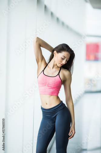 Young beautiful young woman in sportswear doing stretching while standing in front of window at gym
