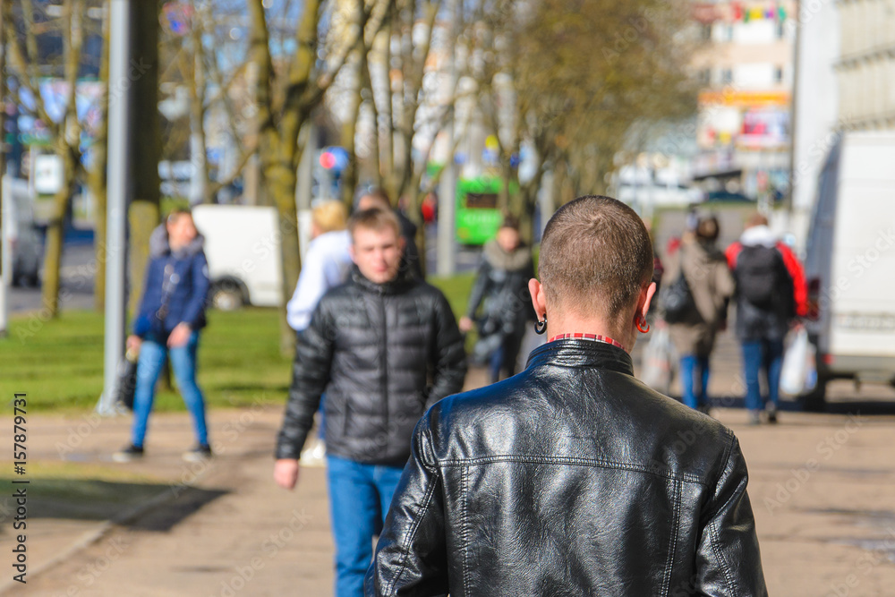 A young man with a short hairstyle and colored earrings in a black leather jacket walks away. View from the back A young man with a short hairstyle and colored earrings in a black leather jacket walks away. View from the back