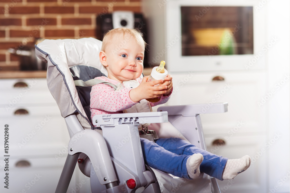 One year toddler girl sits on baby high chair with feeding bottle in her hand. One year toddler girl sits on baby high chair with feeding bottle in her hand.