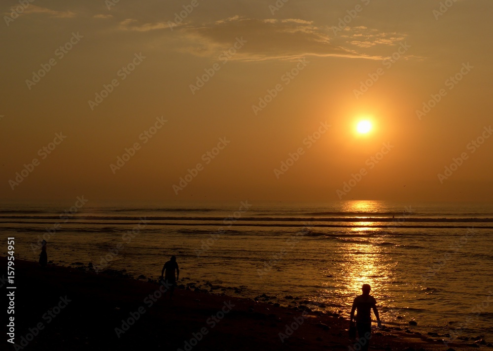 People walking along a beach silhouetted in front of the bright orange glow of the setting sun over the pacific ocean. 