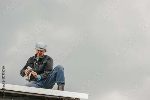 A man in work clothes tighten screws with a screwdriver on the roof against a cloudy sky.