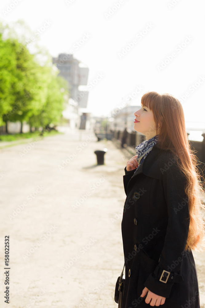 Beautiful red-haired young woman walking down the embankment on a sunny day, examining the neighborhood.