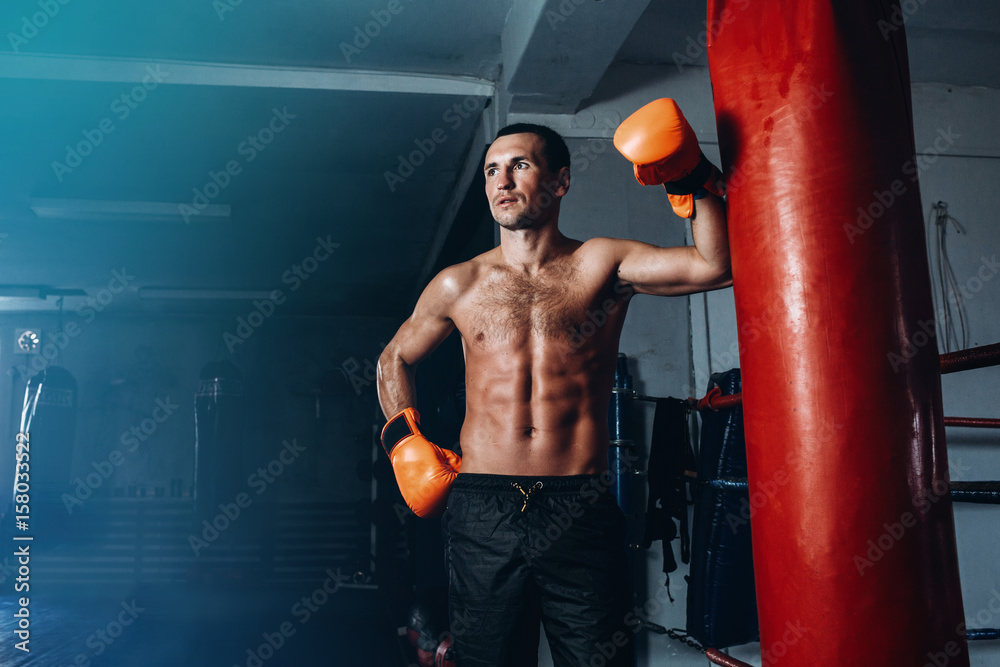 Male boxer training in dark gym. Strong Athletic Man Fitness Model Torso showing six pack abs. Portrait of boxer with gloves