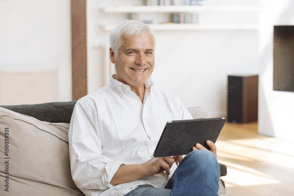Smiling mature man sitting on a sofa using a tablet pc in a living room Smiling mature man sitting on a sofa using a tablet pc in a living room