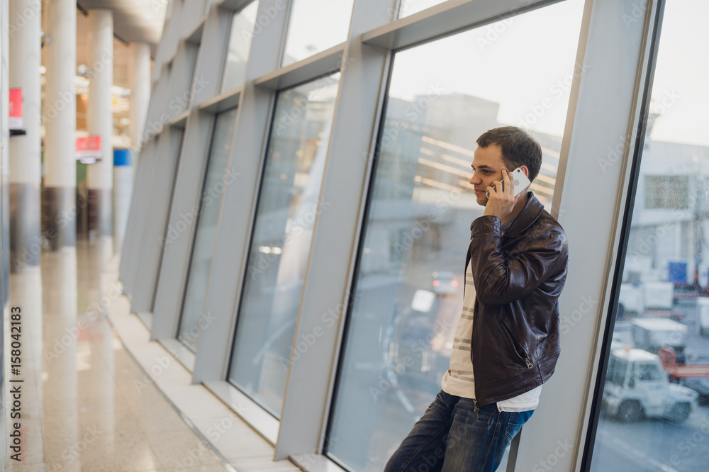 Portrait of young handsome person wearing casual style clothes standing near window in modern airport terminal. Traveler making call using smartphone. Portrait of young handsome person wearing casual style clothes standing near window in modern airport terminal. Traveler making call using smartphone.