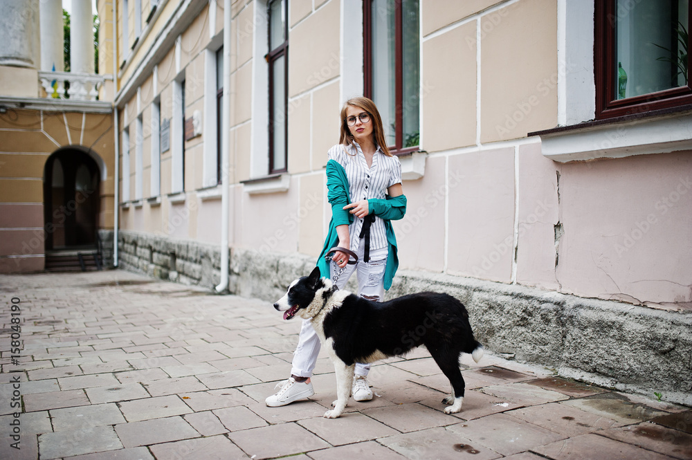 Trendy girl at glasses and ripped jeans with russo-european laika (husky) dog on a leash, against street of city. Friend human with animal theme.