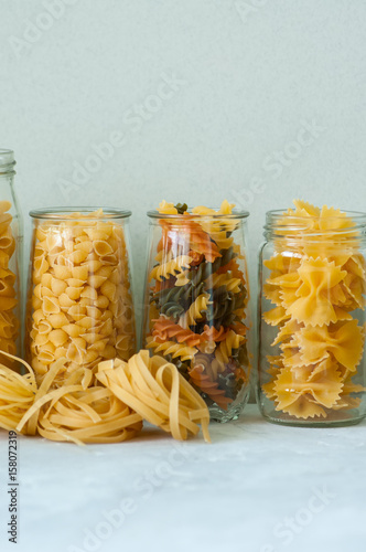 Assortment of different shapes whole grain raw Italian pasta in a glass jar. White stone background. Copy space. Cooking concept.