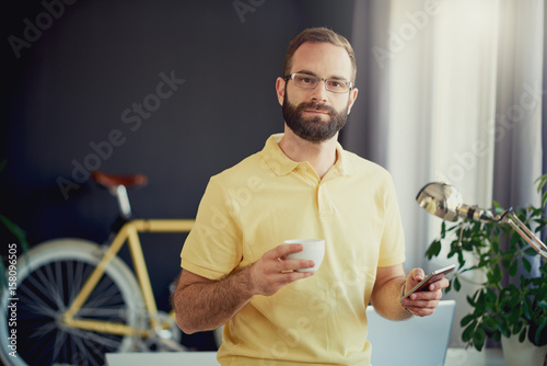 Caucasian freelancer using smartphone and drinking coffee while standing in modern office