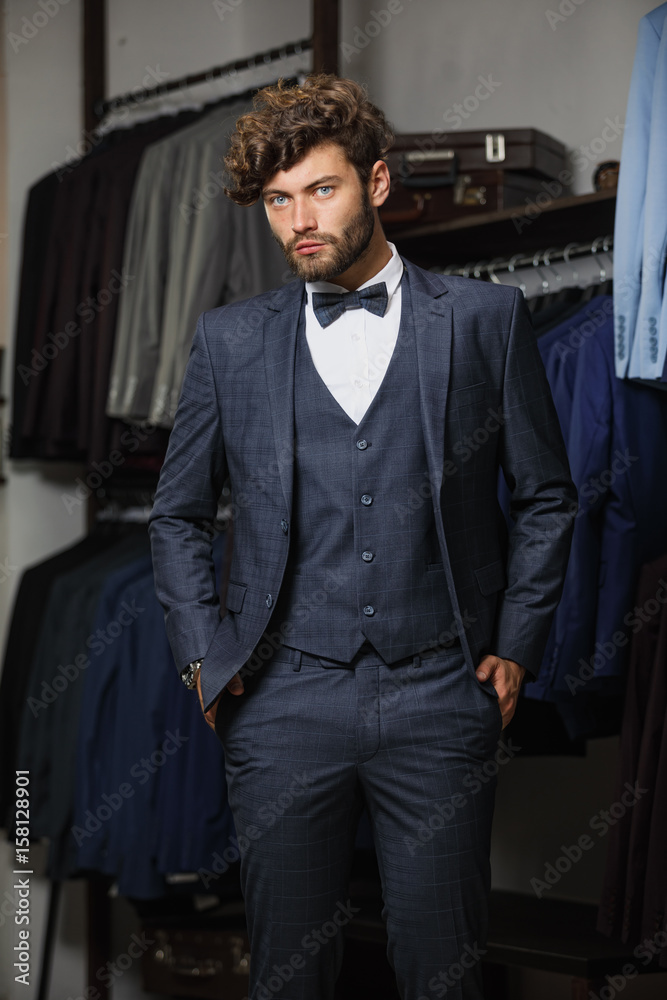 Full length portrait of bearded man in suit standing with crossed arms near the rack with suits in a shop while looking at the camera