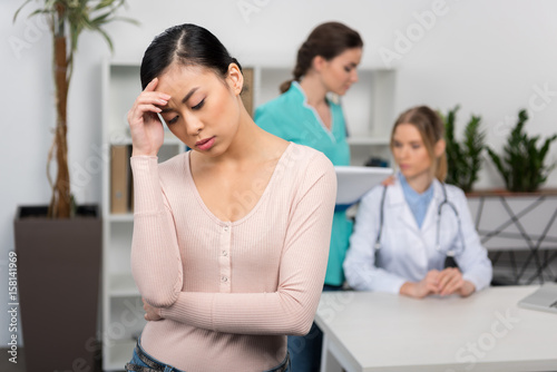 Upset young patient standing with hand on forehead and medical staff talking behind