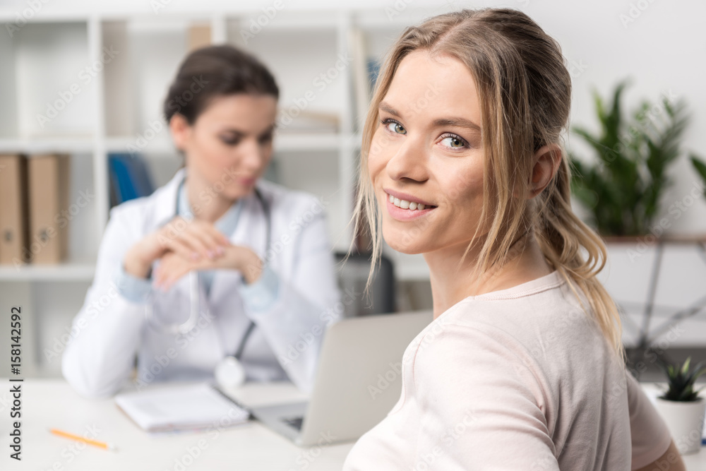 Close-up view of beautiful young patient smiling at camera and doctor using laptop behind Close-up view of beautiful young patient smiling at camera and doctor using laptop behind