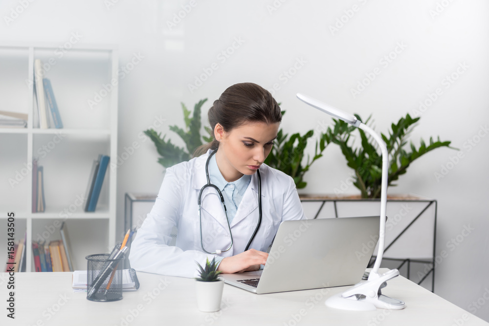 Concentrated young doctor in white coat with stethoscope sitting at table and using laptop in medical office Concentrated young doctor in white coat with stethoscope sitting at table and using laptop in medical office