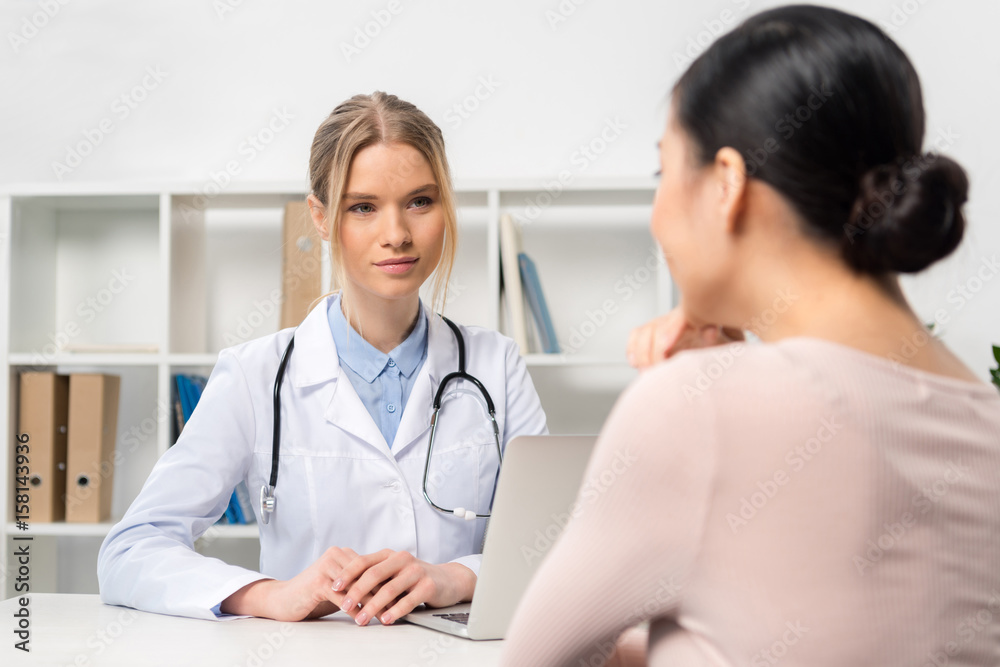 Smiling young doctor with stethoscope sitting at table with laptop and talking with patient Smiling young doctor with stethoscope sitting at table with laptop and talking with patient
