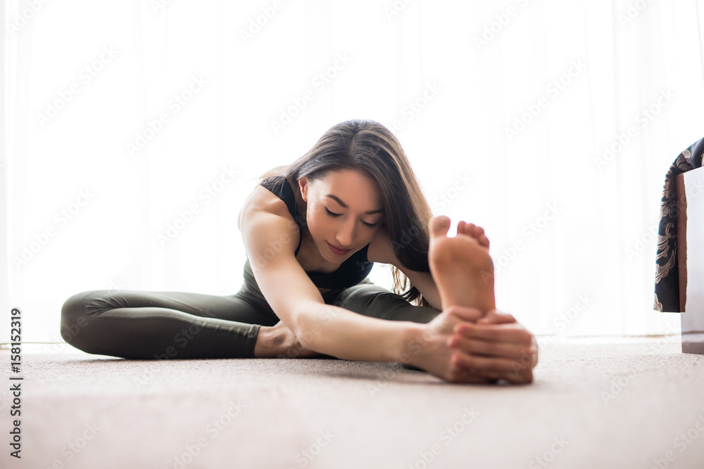 Beautiful young woman in sportswear is smiling while stretching on floor at home