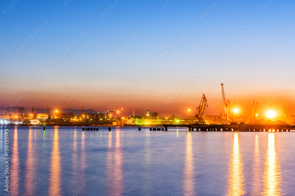 Type of night port. Working freight cranes, a ship at the pier, work in the port. Night photo view of the loading. Beautiful reflection of lights in the sea water.
