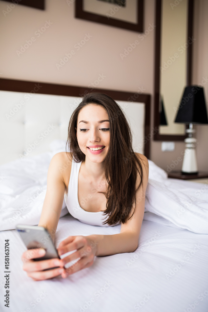Beautiful young woman smiling and holding smart phone while lying in the bed at home Beautiful young woman smiling and holding smart phone while lying in the bed at home