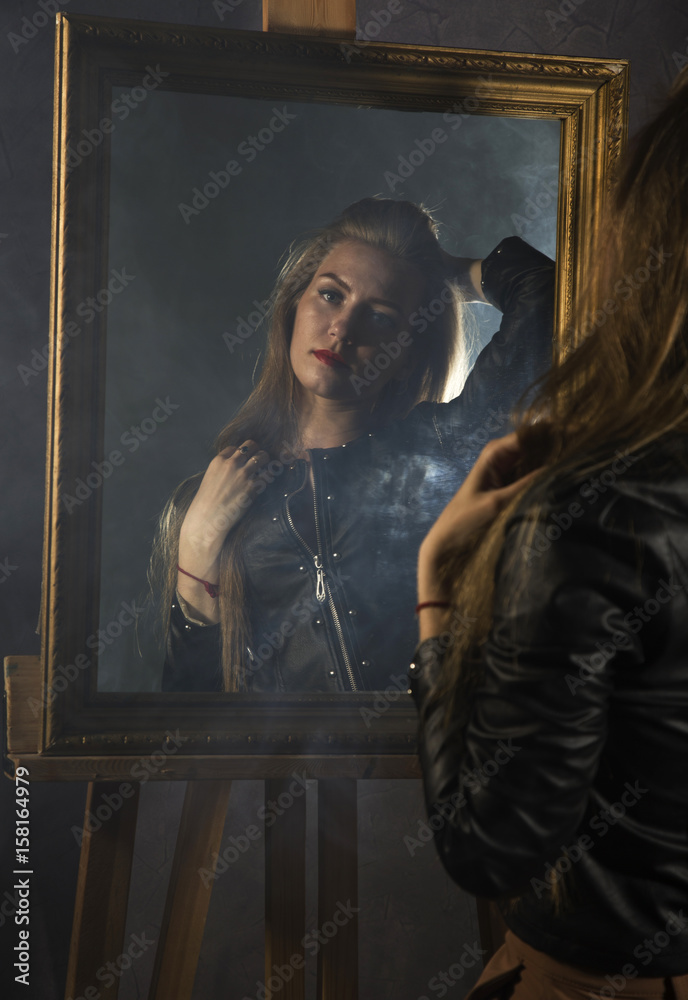 beautiful young woman in a leather jacket looks in the mirror as her portrait beautiful young woman in a leather jacket looks in the mirror as her portrait