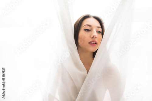 Beautiful young woman looking from behind a white curtain at home in the morning, Happy mood