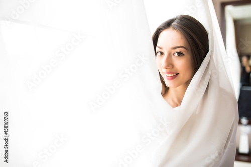 Beautiful young woman looking from behind a white curtain at home in the morning, Happy mood
