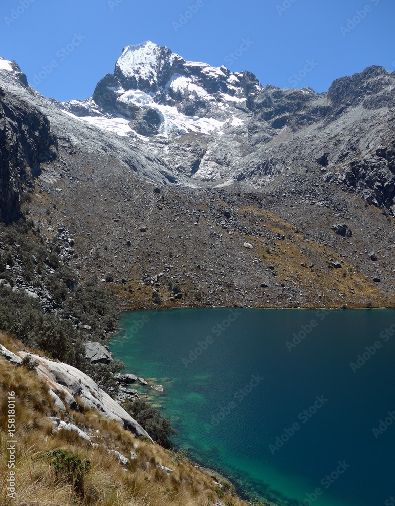 A thin hiking trail passes through the steep rocky hill side above a deep blue/green alpine lake. 