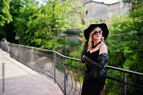 Blonde woman on black dress, leather jacket, sunglasses, necklaces and hat against railings.