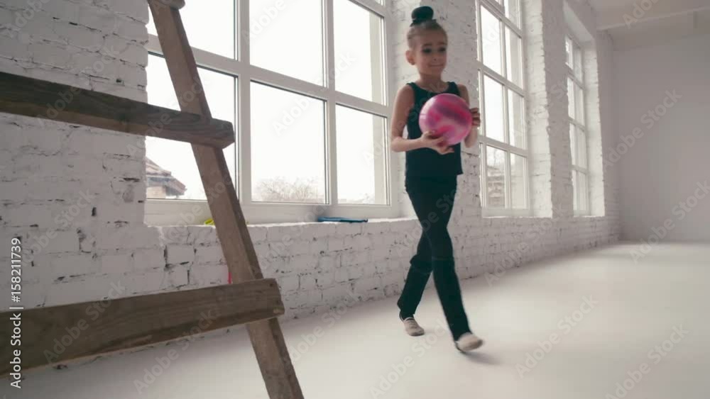 Little girl by the window in a modern specious ballet school prepares to the training, puts on the stocking on her legs, takes the pink ball and goes to the coach. Having fun, active lifestyle.