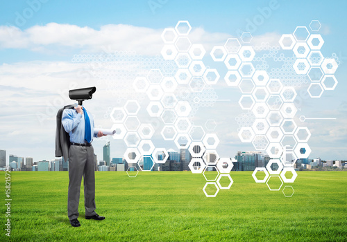 Camera headed man standing on green grass against modern cityscape