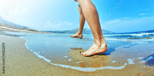 Beach travel - man walking on sand beach on the sand. Closeup detail of male feet and golden sand on beach, Crete, Greece