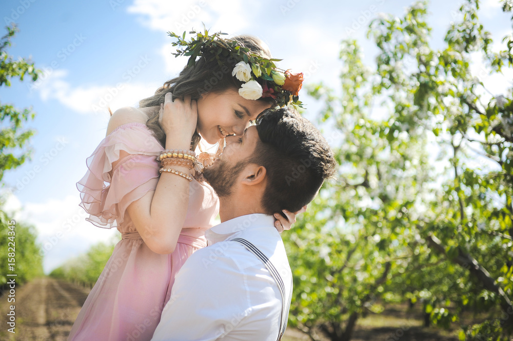 Obraz premium portrait of a girl and couples looking for a wedding dress, a pink dress flying with a wreath of flowers on her head on a background garden and the blue sky, and they hug and pose 