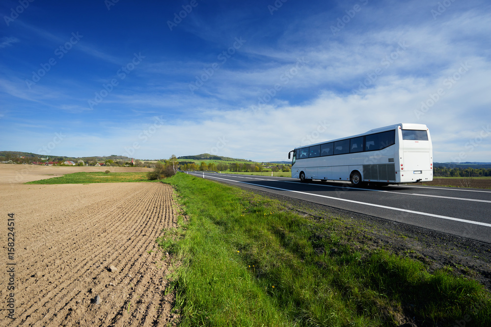 The white bus traveling on the road around a sown field in a rural landscape with a village in the background The white bus traveling on the road around a sown field in a rural landscape with a village in the background