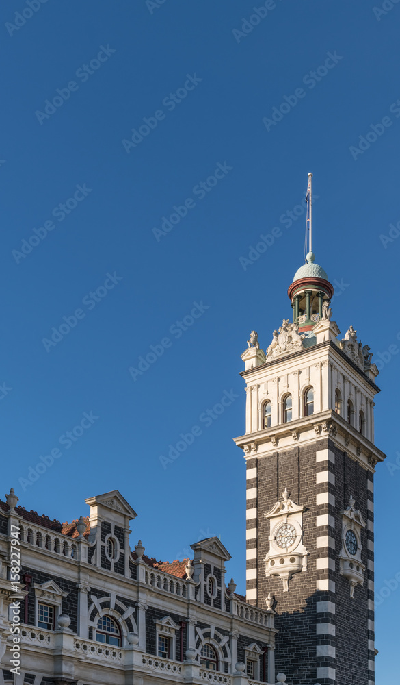 Dunedin, New Zealand - March 15, 2017: Clock tower and part of facade in brown and cream stones of historic railway station against full blue sky.