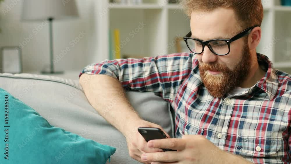 Attractive man with smartphone sitting on sofa at home in the living room and chatting with friends.