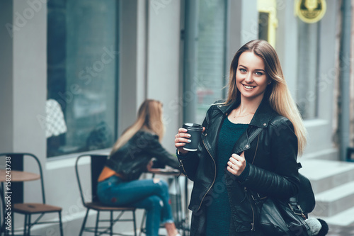 Take away coffee. Beautiful young urban woman wearing in black stylish clothes holding coffee cup and smiling while walking along the street. Student's coffee break after study. Fashion lifestyle.