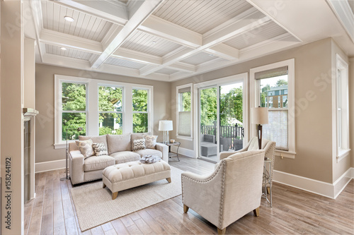 Beautiful living room with hardwood floors in new luxury home. Coffered ceiling adds elegant touch to this furnished living space. Lush trees provide scenic exterior view. 