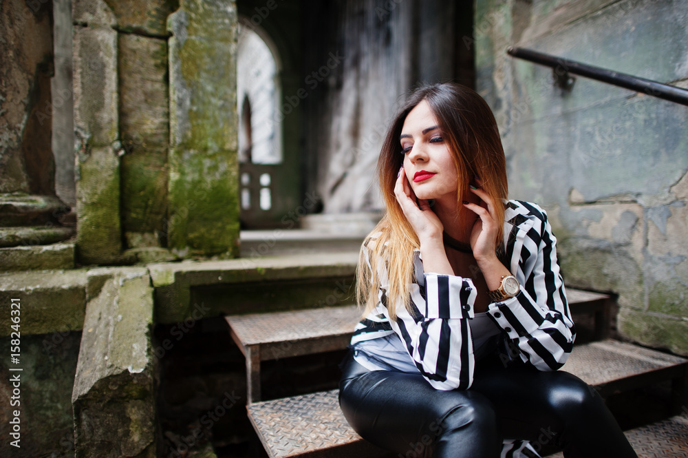 Fototapeta premium Fashionable woman look with black and white striped suit jacket, leather pants, posing at old street on iron stairs. Concept of fashion girl.