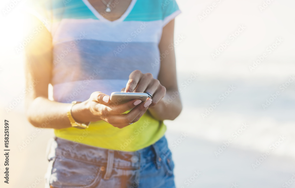 Hipster girl on smart phone gadget in sand coastline, mock up of blank screen. Traveler hold and using in female hand mobile on background seascape horizon. Tourist look on blue sun ocean, lifestyle