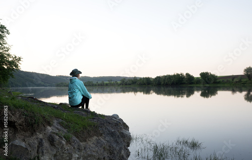 Female traveller sits on the stone above the river. Sunset time. Hills and forest on the banks