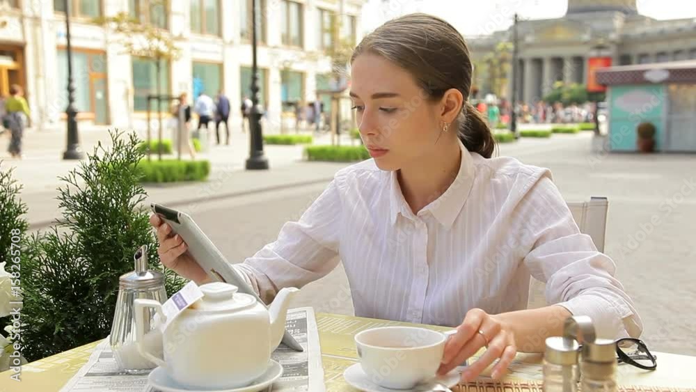 Female caucasian student in outdoor cafe with tablet. Drinks coffee