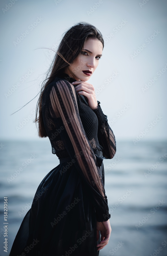 Beautiful sad goth girl in black dress standing on sea beach Beautiful sad goth girl in black dress standing on sea beach