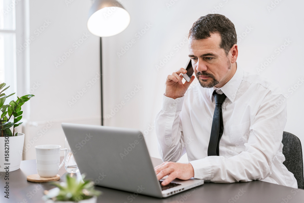 Middle aged man making call and using his laptop while working from home office Middle aged man making call and using his laptop while working from home office