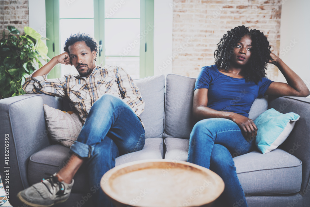 Young displeased black couple.American african men arguing with his stylish  girlfriend,who is sitting on sofa on couch next to him with legs  crossed.Man looking away offended expression on her face. Stock Photo