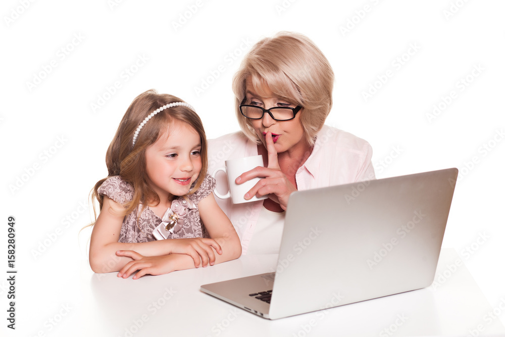 Grandmother and her granddaughter sitting at the table and using laptop on white background Grandmother and her granddaughter sitting at the table and using laptop on white background