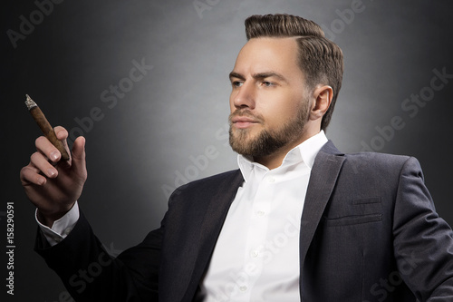 Bearded man holding cigar in hand. Formal wear in suit and shirt