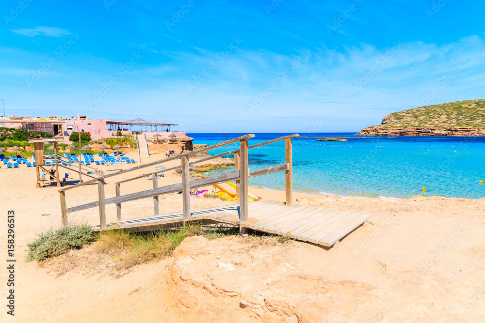 Wooden walkway on sandy Cala Comte beach and restaurant building on shore, Ibiza island, Spain.