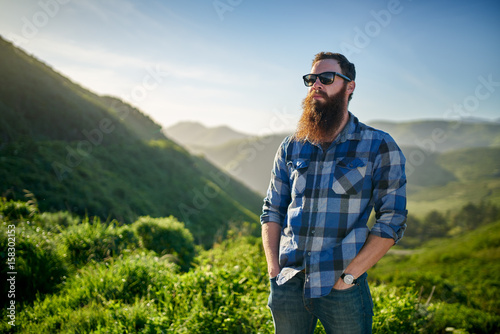 bearded guy with sunglasses and blue flannel posing in front of grassy green hills in california photo